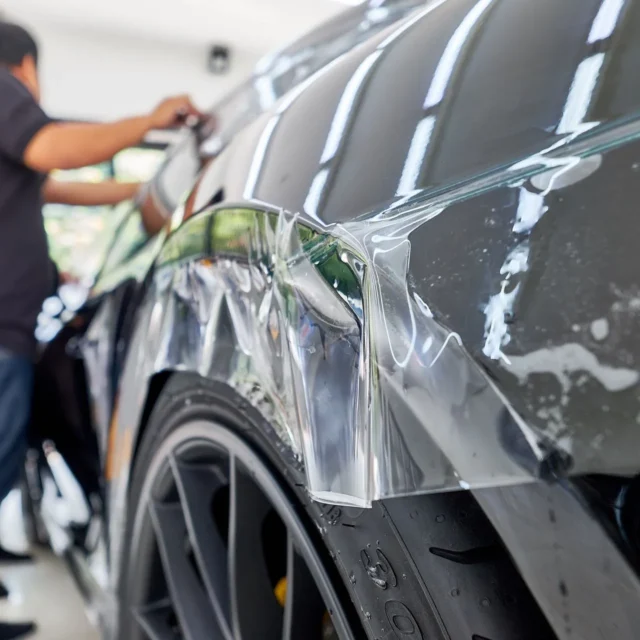 Installer fitting clear paint protection film over a black car’s rear quarter panel around the wheel arch.